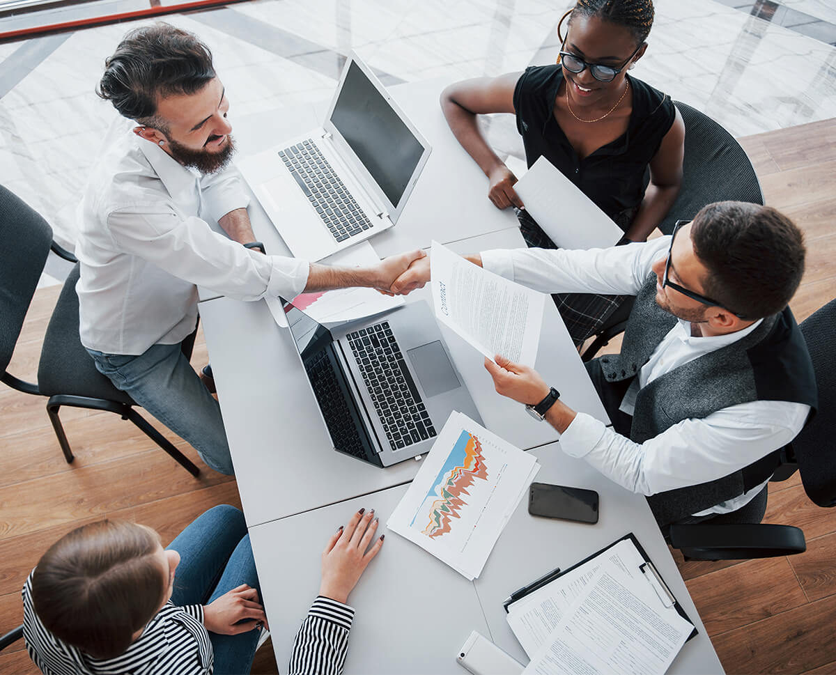 4 mensen rondom een tafel met computers op de tafel in een zakelijke setting, twee personen schudden handen.